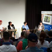 Fünf Personen sitzen an Tischen auf einer Bühne. Vier haben ihren Blick der fünften sprechenden Person zugewendet, sie sitzt am äusseren rechten Rand. Im Vordergrund einige Köpfe aus dem Publikum.