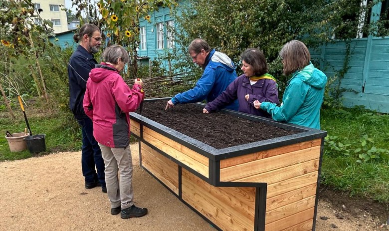 Mehrere Personen stehen um ein rollstuhlgerechtes Hochbeet. Eine längsseite davon ist "eingebuchtet" und bietet Platz für Stuhl und Beine.