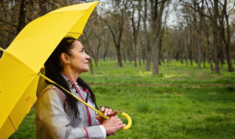 Eine Frau mit einem gelben Regenschirm steht auf einer Wiese und blickt zu den Bäumen im Hintergrund.